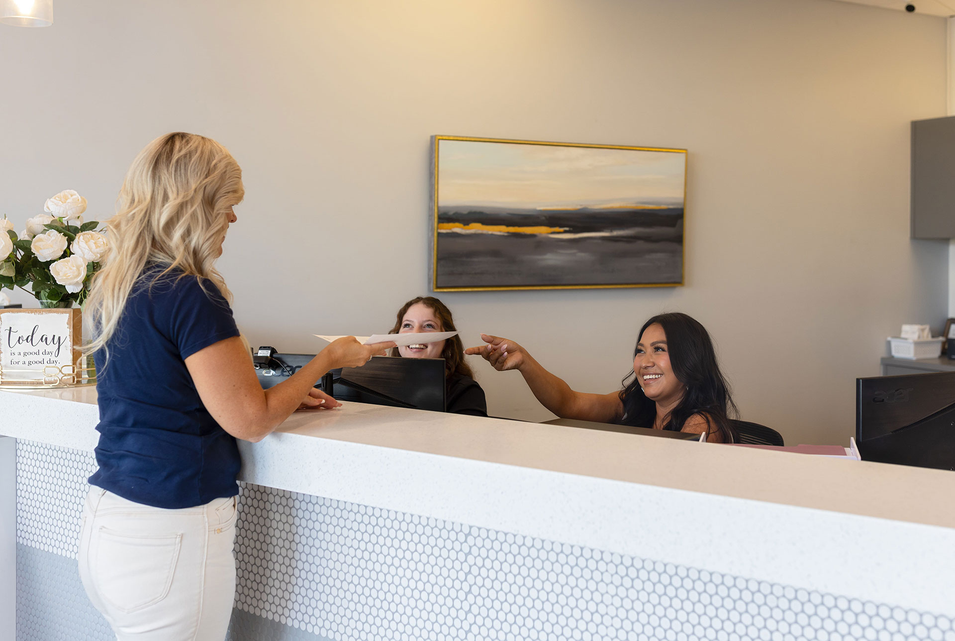 In the image, two women are interacting at a counter with a receptionist. One woman is handing something to the other who appears to be receiving it. The setting suggests an office environment.