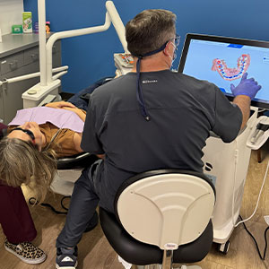 A dentist examining a patient s mouth using a dental chair with adjustable height and a computer screen displaying 3D imagery of the teeth.